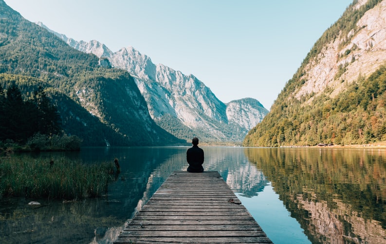 Man sitting on wooden dock facing a lake and mountains