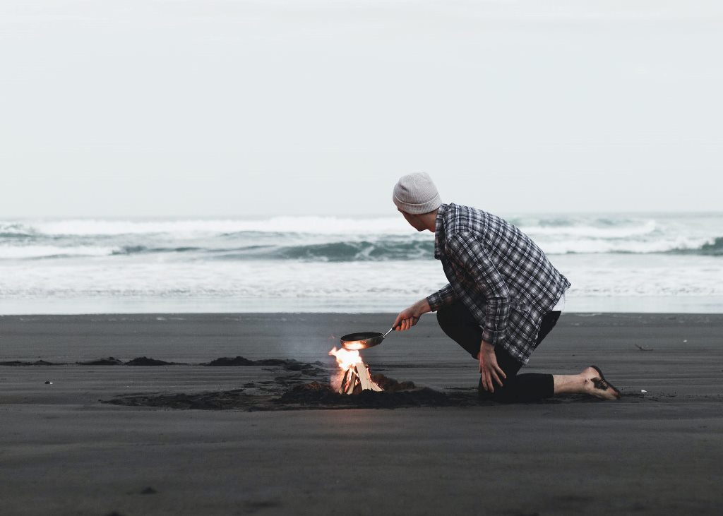 A man cooking in a small campfire along the seashore